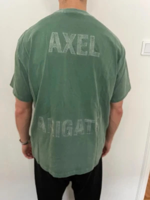 Grön t-shirt från Axel Arigato - Säljer en grön t-shirt från Axel Arigato med tryck på ryggen. T-shirten har en relaxed passform, rund hals och korta ärmar. 