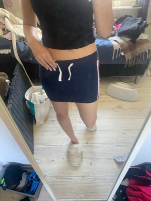 Mörkblå kortkjol med snörning - Sportig mörkblå kortkjol med vita och röda ränder på sidorna och vit snörning i midjan. Kjolen har en tight passform och är perfekt för en avslappnad och trendig look.