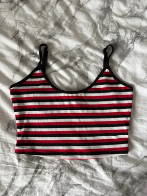 Randig croppad linnetopp - Säljer en croppad linnetopp med smala axelband och ribbad struktur. Toppen har ett randigt mönster i svart, rött och vitt med svarta kanter. Perfekt för varma dagar och enkel att styla till jeans eller kjol.