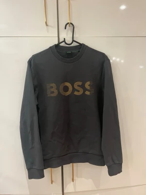 Mörkgrå sweatshirt från BOSS - Snygg mörkgrå sweatshirt från BOSS med guldfärgat logotryck på bröstet. Tröjan har rund halsringning, ribbade muddar och en avslappnad passform. Perfekt för en trendig och casual look. Skriv för mer information eller information 