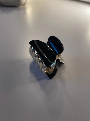 Svart hårklämma - Snygg hårklämma i svart och silver med stilren design. Klämman har en rektangulär form och metalliska detaljer som ger en modern touch. Perfekt för att snabbt sätta upp håret och addera en trendig accessoar till din look. Aldrig använd