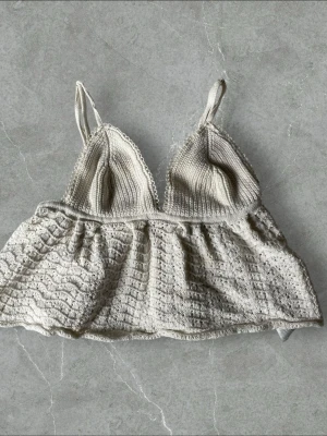 Vit virkad topp med tunna band - Söt vit virkad topp med tunna axelband och spetsdetaljer. Toppen har en mjuk, vågig kant och knytband i ryggen för justerbar passform. Materialet är virkat och ger en bohemisk känsla. Perfekt för dig som gillar romantisk och avslappnad stil.