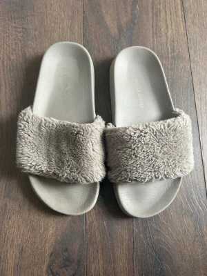  fluffiga slip-in tofflor - Säljer ett par gråa slip-in tofflor med breda, mjuka band i fluffigt fuskpälsmaterial. Sulan är matchande och har en enkel, stilren design. Perfekta för att chilla hemma.
