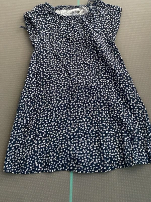 Mörkblå blommig kortklänning till den mindre - Söt mörkblå kortklänning med vitt blommönster och rund hals. Klänningen är ärmlös och har en avslappnad passform, perfekt för varma dagar. Tillverkad i ett lätt och mjukt material som känns skönt mot huden.