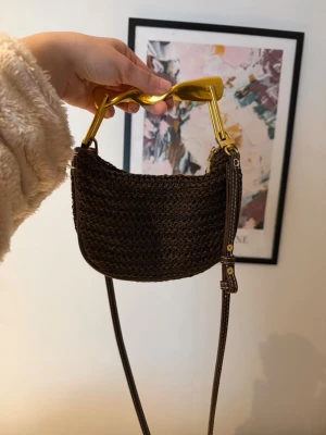 Brun virkad handväska med gulddetaljer - Snygg brun virkad handväska med unik guldfärgad handtag och lång axelrem i läderimitation. Väskan har en halvmåneformad design och är perfekt för dig som vill ha något trendigt och annorlunda. Rymmer det viktigaste och ger din outfit en cool touch.