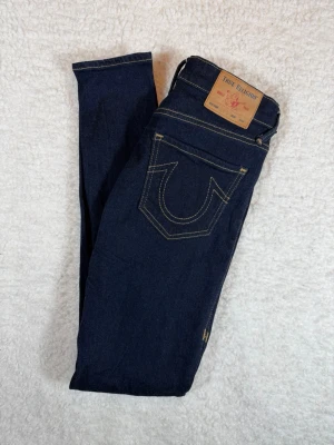 Mörkblå skinny jeans True Religion - Snygga mörkblå skinny jeans från True Religion med klassisk kontrastsöm och ikonisk bakficka med U-formad brodyr. Jeansen har fem fickor, knapp- och dragkedjegylf samt läderpatch bak. Perfekt för dig som gillar en tight och modern look.