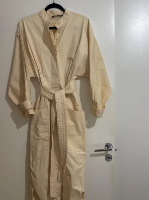 Beige långklänning med bälte - Stilren beige långklänning med knappar framtill, stora fickor och ett matchande bälte i midjan. Klänningen har lång ärm och upprullade ärmslut för en avslappnad look. Perfekt för dig som gillar minimalistisk och enkel stil.