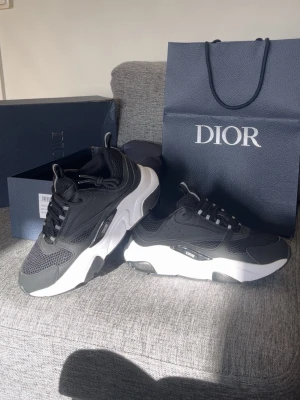 Svarta Dior sneakers med vit sula - Snygga svarta Dior sneakers med chunky vit sula och meshdetaljer. Skorna har svarta och vita snören, samt diskret Dior-logga på sidan. Kombination av mesh och läder ger en modern och sportig look. Perfekta för dig som vill ha en exklusiv touch på din outfit.
