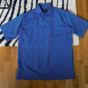 Blårutig kortärmad skjorta Adolfo - Snygg blårutig skjorta från Adolfo med korta ärmar och klassisk krage. Skjortan har knappar framtill och är tillverkad i ett lätt bomullsmaterial som passar perfekt till sommaren. Enkel och stilren design som funkar till många olika looks.