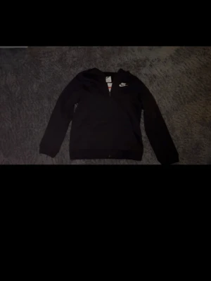 Nike zip hoodie - Säljer en svart hoodie från Nike med dragkedja framtill och broderad vit Nike-logga på bröstet. Hoodien har långa ärmar och ribbade muddar vid ärmslut och nederkant. Perfekt för en sportig och avslappnad stil.