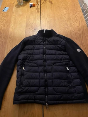 Svart pufferjacka från Moncler - Snygg svart pufferjacka från Moncler med quiltad front och stickade ärmar. Jackan har två dragkedjeförsedda fickor, hög krage och Moncler-logga på ärmen. Perfekt för kalla dagar och har en modern, sportig look. Går att scanna NFC storlek L men passar M