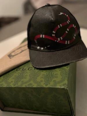 Svart Gucci keps med ormtryck - Säljer en svart keps från Gucci med ett fett ormtryck i rött, vitt och svart på framsidan. Kepsen har böjd skärm och mesh på baksidan för extra ventilation. Justerbar rem bak för perfekt passform. Riktigt snygg accessoar för dig som vill sticka ut.