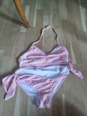 Pastellfärgad bikini från Kappahl - Säljer en snygg bikini från Kappahl i pastelliga rosa, lila och beige toner. Underden har knyt i sidorna och ett swirl-mönster som ger en retrovibe. Överdelen är halterneck och blir lite smalare på baksidan.
