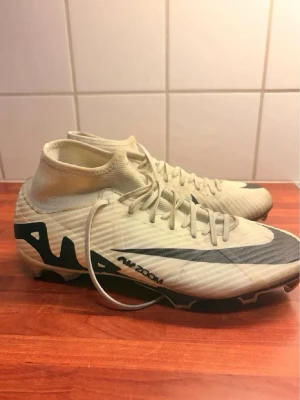 Nike Air Zoom vita fotbollsskor - Säljer ett par vita Nike Air Zoom fotbollsskor med högt skaft och svarta detaljer. Skorna har snörning, en svart Nike-swoosh på sidan och mönstrad sula för bra grepp på planen. Tillverkade i syntetmaterial för lätt känsla och bra bollkontroll.