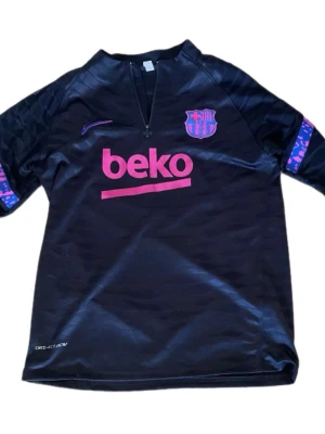 Barcelona träningsset svart/lila Nike - Säljer ett svart Barcelona träningsset från Nike med lila och rosa detaljer. Tröjan har halv dragkedja, klubbmärke och sponsortryck framtill. Byxorna har elastisk midja, klubbmärke och färgglada band längs benen. Perfekt för fotbollsträning eller chill.