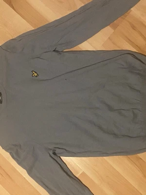 Grå tröja från Lyle & Scott - Snygg grå tröja från Lyle & Scott med klassisk gul logga på bröstet. Tröjan har rund halsringning, långa ärmar och ribbade muddar vid ärmslut och nederkant. Perfekt för en stilren och enkel look. Storlek M men funkar som S