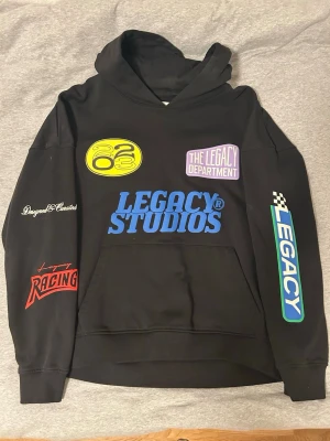 Legacy Studios svart hoodie med tryck - Svart hoodie från Legacy Studios med flera färgglada tryck på framsidan, perfekt för streetwear. Jättesnygg och skön endast använd fåtal gånger!