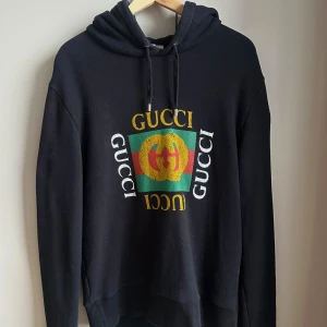 Svart hoodie från Gucci med tryck - Säljer en svart hoodie från Gucci med färgglatt tryck på bröstet och logga i gult, grönt och rött. Klassisk huva med dragsko och ribbade muddar. Tillverkad i Italien. Perfekt för dig som vill ha en statement-piece i garderoben.