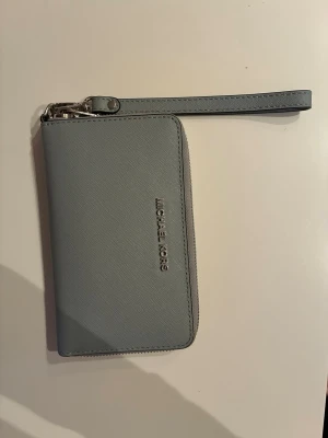 Ljusgrå plånbok från Michael Kors - Snygg ljusgrå plånbok/wallet från Michael Kors. Plånboken har dragkedja runtom, handledsrem, flera kortfack, myntfack med dragkedja och ett fönster för ID eller busskort. Silver detaljer. Ser ut som saffiano läder. Använd ett fåtal ggr. Är i mkt bra skick, inga defekter. 17x10 cm
