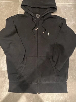 Svart hoodie från Polo Ralph Lauren - Klassisk svart hoodie från Polo Ralph Lauren med dragkedja framtill, två fickor och justerbar huva. Ikoniska vita logobrodyr på bröstet. Tillverkad i mjuk bomullsblandning som är perfekt för en avslappnad och stilren look.