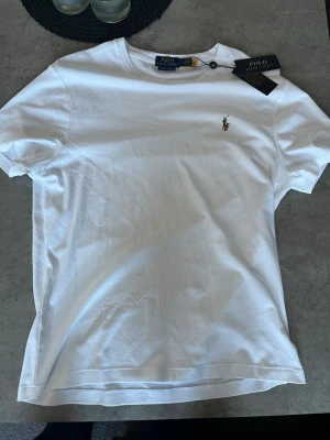 Vit t-shirt från Polo Ralph Lauren - Klassisk vit t-shirt från Polo Ralph Lauren med broderad logga på bröstet. Modellen är custom slim fit och har rund hals samt korta ärmar. Tillverkad i mjuk bomull för en skön känsla och stilren look.