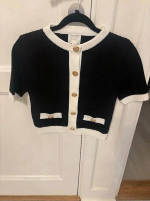 Svart kort kofta med vita detaljer - Trendig svart kort kofta med vita ribbade kanter och guldknappar framtill. Koftan har korta ärmar och två dekorativa fickor med vita rosetter. Perfekt för att lyfta en enkel outfit med en chic touch.