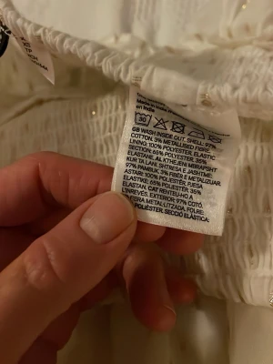 Vit volangkjol med guldprickar XS - Supersöt vit kjol med flera volanger och små guldprickar. Kjolen har bred smock i midjan för skön passform och är gjord av bomull med metalliserad fiber. Perfekt för dig som gillar en lekfull och trendig stil.