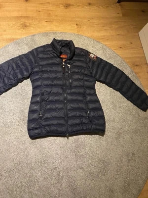 dunjacka från Parajumpers - Snygg mörkblå dunjacka från Parajumpers med quiltad design, hög krage och dragkedja framtill. Jackan har tre fickor med dragkedja och en patch med logga på ena ärmen. Perfekt för kalla vinterdagar och riktigt lätt att bära. Storleken är L men sitter som en storlek S. Billigare vid snabb affär 