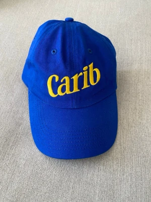 Blå Carib keps med gul brodyr - Snygg blå keps från Carib med justerbar rem bak och gul broderad logga framtill. Klassisk böjd skärm och ventilerande hål upptill. Tillverkad i bomullstyg som känns lätt och bekvämt på huvudet. Perfekt för dig som vill sticka ut med färg och stil.
