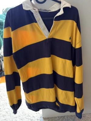 Randig rugbytröja från Ralph Lauren - Klassisk rugbytröja med breda marinblå och gula ränder, vit krage och knappslå. Långärmad modell med avslappnad passform och broderad logga på bröstet. Perfekt för en sportig och stilren look.