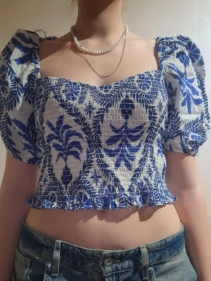 Blåvit croppad blus med puffärm - Trendig croppad blus med blått och vitt blommönster, puffiga korta ärmar och smockad kropp. Fyrkantig ringning och knyt i ryggen ger en söt och somrig vibe. Perfekt till höga jeans eller kjol. (Aldrig använd, fel storlek)