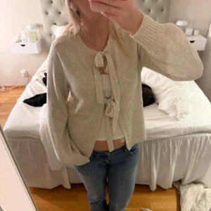 Beige kofta med knytband - Säljer en beige kofta med två dekorativa knytband framtill. Koftan har lång ärm, ribbade muddar och en mjuk, stickad känsla. Perfekt att slänga över en topp för en avslappnad men söt look.