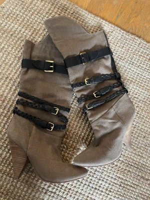 Bruna stövlar med svarta remmar - Snygga bruna stövlar med höga klackar och spetsig tå. Dekorerade med svarta bälten och flätade remmar med guldfärgade spännen runt skaftet. Tillverkade i textil med träliknande klack. Några lösa trådar men det kan man klippa bort 