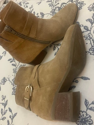 Beige mocka stövletter med klack - Snygga beige stövletter i mocka med bred klack och rundad tå. Dragkedja på insidan och dekorativt spänne i guld på sidan. Perfekta för dig som vill ha en stilren och trendig look.