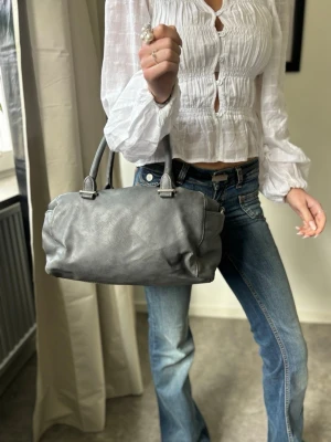 Grå handväska i skinn med dubbla handtag - Säljer en snygg grå handväska i skinn med dubbla handtag och silverfärgade detaljer. Väskan har en mjuk, rundad form och stängs med dragkedja. Perfekt storlek för det viktigaste och passar till många olika outfits.