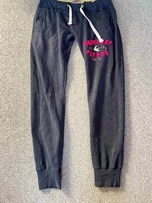 Mörkblå mjukisbyxor Superdry - Snygga mörkblå mjukisbyxor från Superdry med vita dragsnören och rosa tryck på ena benet där det står 'Superdry Players Tokyo'. Byxorna har muddar vid bensluten och två fickor fram. Storlek XS
