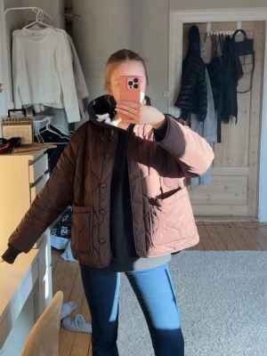 Brun quiltad vindjacka med knappar - Snygg brun quiltad vindjacka med knäppning framtill och två stora fickor. Jackan har en boxy passform och vågigt mönster i tyget. Perfekt för lager-på-lager och ger en avslappnad men trendig look.