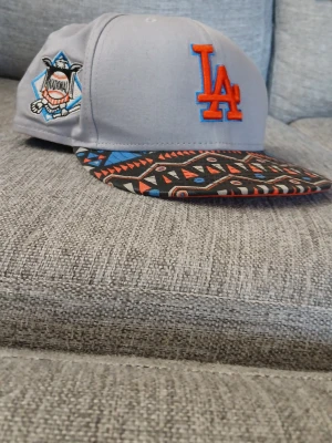 Grå LA-keps med mönstrad skärm - Säljer en grå snapback-keps från New Era med LA Dodgers-logga i orange och blått framtill. Skärmen har ett färgstarkt, geometriskt mönster i svart, orange, blått och beige. Sidan pryds av en broderad patch och New Era-logga i orange.