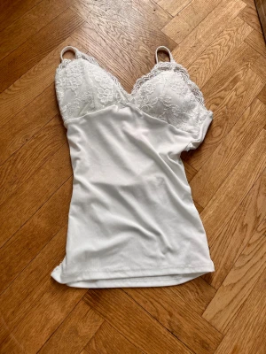 Vit spetsdetaljerad topp med smala band - Säljer en vit topp med tunna axelband och spetsdetaljer över bysten. Toppen har en mjuk och stretchig känsla, med slät tyg över magen och vacker blommig spets upptill. Perfekt för lager på lager eller som ett statement-plagg.Skriv gärna för mått! 💕