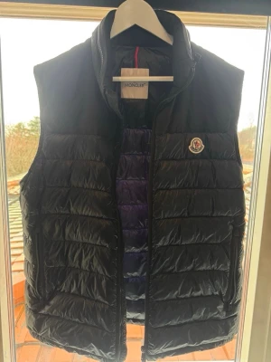 Mörkblå dunväst från Moncler - Snygg mörkblå dunväst från Moncler med klassisk quiltad design och hög krage. Västen har Moncler-logga på bröstet och dragkedja framtill. Perfekt för lager-på-lager och håller dig varm med lätt dunfyllning.
