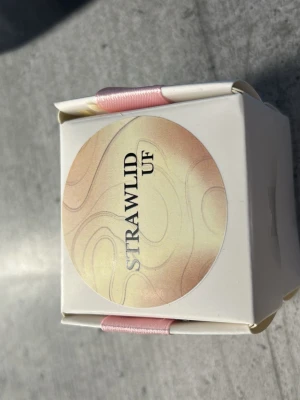 StrawLid UF förpackning - En liten vit förpackning med rund etikett i beige och rosa toner där det står 'STRAWLID UF'. Förpackningen är dekorerad med ett rosa band och har en stilren, modern design. Perfekt för dig som gillar smarta tillbehör.