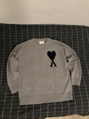 Grå stickad tröja från Ami Paris - Säljer en grå stickad tröja från Ami Paris med svart hjärta och A-logga på bröstet. Tröjan har rund hals, ribbade muddar och är långärmad. Perfekt för lager-på-lager och chill stil. Använt typ 2 gånger. Är i nyskick, hör av dig om du har några frågor.