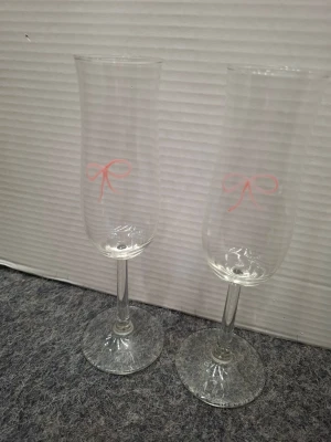 Två champagneglas med rosa rosett - Säljer två eleganta champagneglas i klart glas med en söt rosa rosett tryckt på framsidan. Glasen har klassisk form med smal kupa och stabil fot, perfekta för festliga tillfällen eller som dekoration.