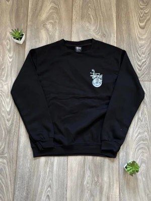 Svart Stüssy sweatshirt med tryck - Svart sweatshirt från Stüssy med vitt tryck på bröstet som visar loggan och en discokula med siffran 8. Tröjan har rund halsringning, långa ärmar och ribbade muddar. Perfekt för en streetwear-look.