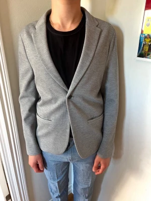 Ljusgrå kavaj - Snygg ljusgrå kavaj med smal passform och enkel knappstängning. Kavajen har två fickor framtill och klassisk krage med slag. Perfekt att styla med en t-shirt för en avslappnad look. Materialet känns mjukt och följsamt.