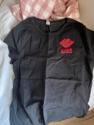 Svart t-shirt med rött KISS-tryck - Svart t-shirt från Only Young med korta ärmar och ett rött tryck av läppar samt texten 'KISS' på bröstet. Tillverkad i mjuk bomull och har en klassisk rund halsringning. Perfekt för dig som vill ha en enkel men ändå uttrycksfull look.