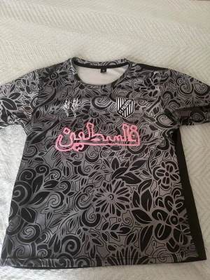 Fotbolls tröja - Unik sporttröja i svart och grått med blommigt mönster och rosa text på bröstet. Tröjan har rund hals, korta ärmar och klubbmärke på vänster sida. Tillverkad i ett lätt och snabbtorkande material, perfekt för träning eller match.