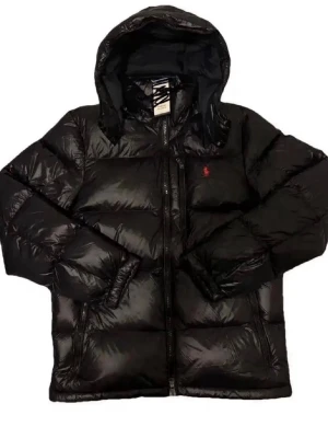 Svart pufferjacka från Polo Ralph Lauren - Snygg svart pufferjacka från Polo Ralph Lauren med huva och dragkedja framtill. Jackan har två sidofickor och den klassiska röda loggan broderad på bröstet. Tillverkad i glansigt material som ger en modern look och håller dig varm under kalla dagar.