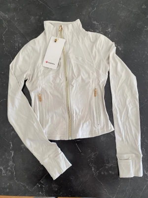 Vit Define Cropped Jacket från Lululemon - Snygg vit cropped träningsjacka från Lululemon i det mjuka och stretchiga Nulu-tyget. Jackan har figurnära passform, tumhål vid ärmslut, två fickor med dragkedja och guldfärgade detaljer. Perfekt för gymmet eller som ett sportigt lager.