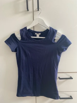 Mörkblå topp med cut out från Amisu - Snygg mörkblå topp från Amisu med trendiga cut out-detaljer vid axlarna. Modellen är figurnära med korta ärmar och rund halsringning. Tillverkad i mjuk bomull som känns skön mot huden. Perfekt för dig som vill ha en enkel men cool look.